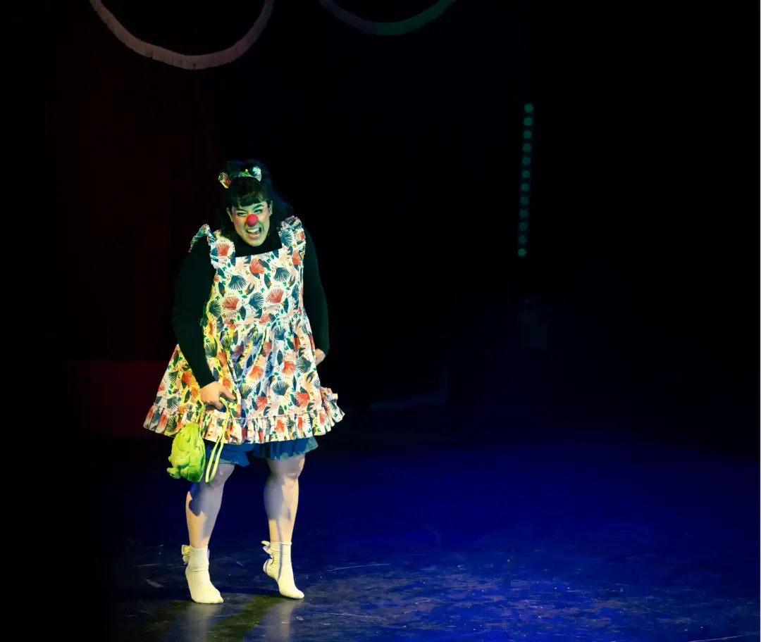 A clown in a floral pinafore grimacing on a dark stage, holding a green bag