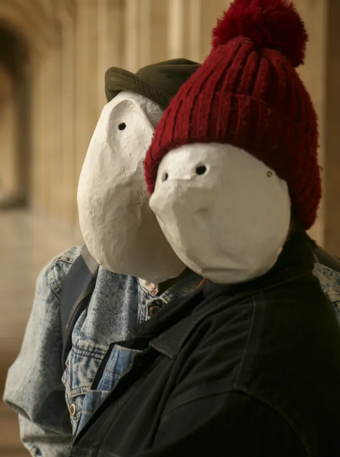 Two performers in white larval masks, one wearing a red knit hat