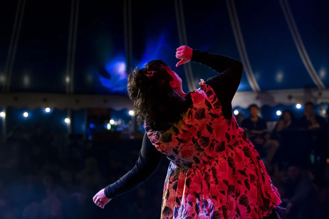 A clown in a red floral dress dancing with arms raised inside a circus tent