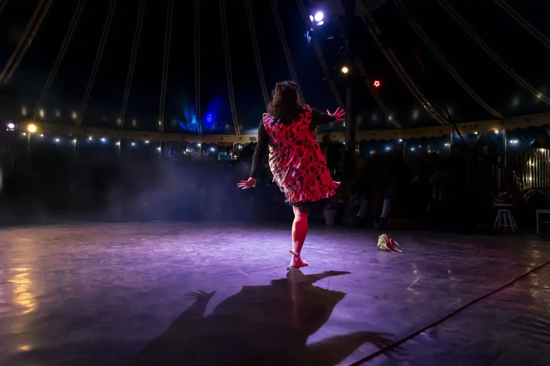 A clown in a red dress on a lit circus stage, casting a long shadow