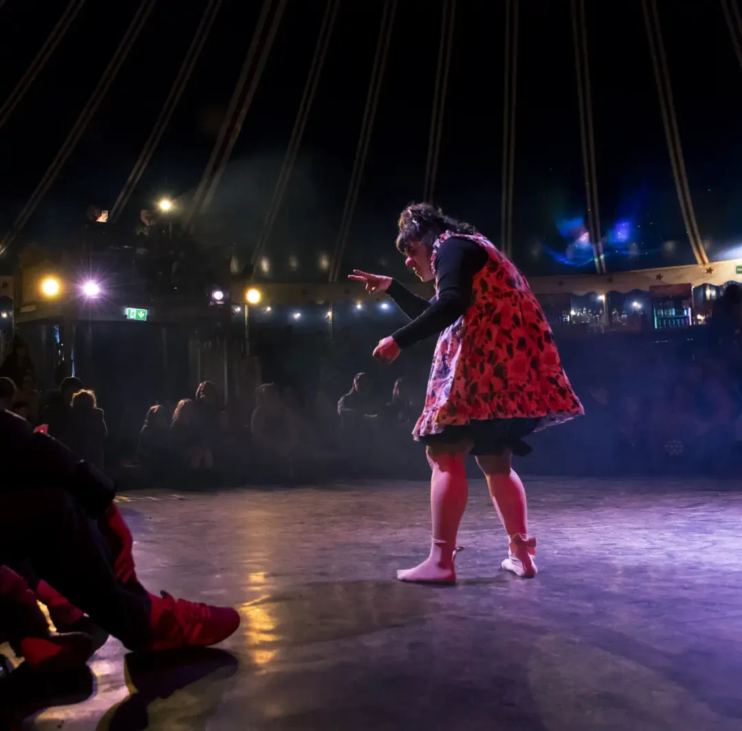 A clown in a red dress bending toward a toy on the circus stage, the audience behind
