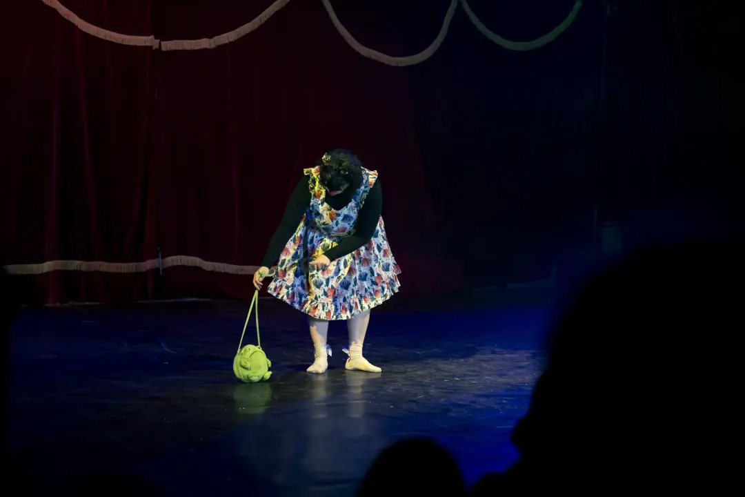 A clown in a floral pinafore on a dark stage, a green bag at her feet