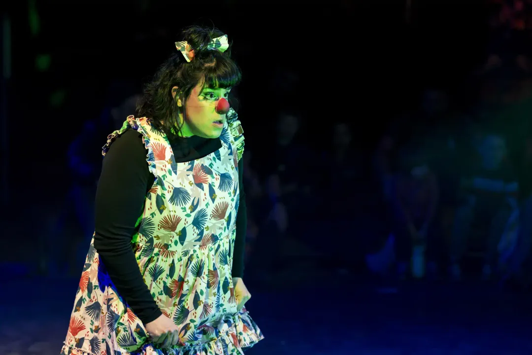 A clown with a red nose in a floral pinafore, glancing to the side